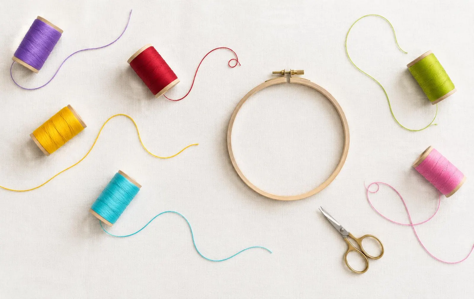 Thread spools in violet, red, yellow, aqua, green and pink around an empty wooden hoop and small scissors on white linen